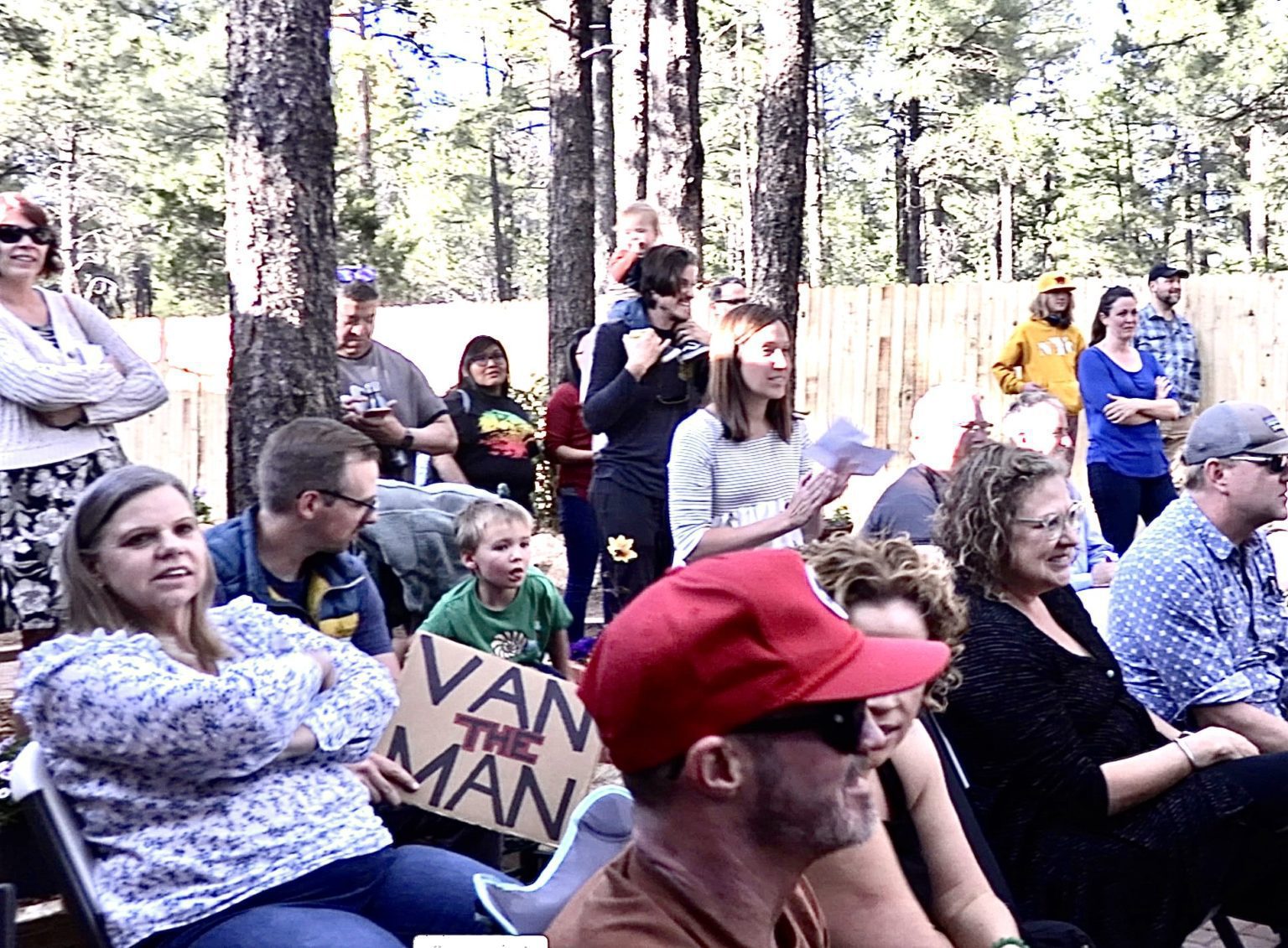 Audience of parents and kids watching an outdoor show among pine trees, one holding a Van the Man sign