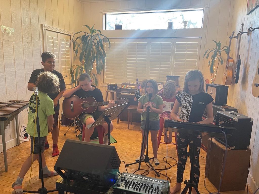 Kids playing acoustic guitar, singing and playing keyboard in a sunny wood-floored music room