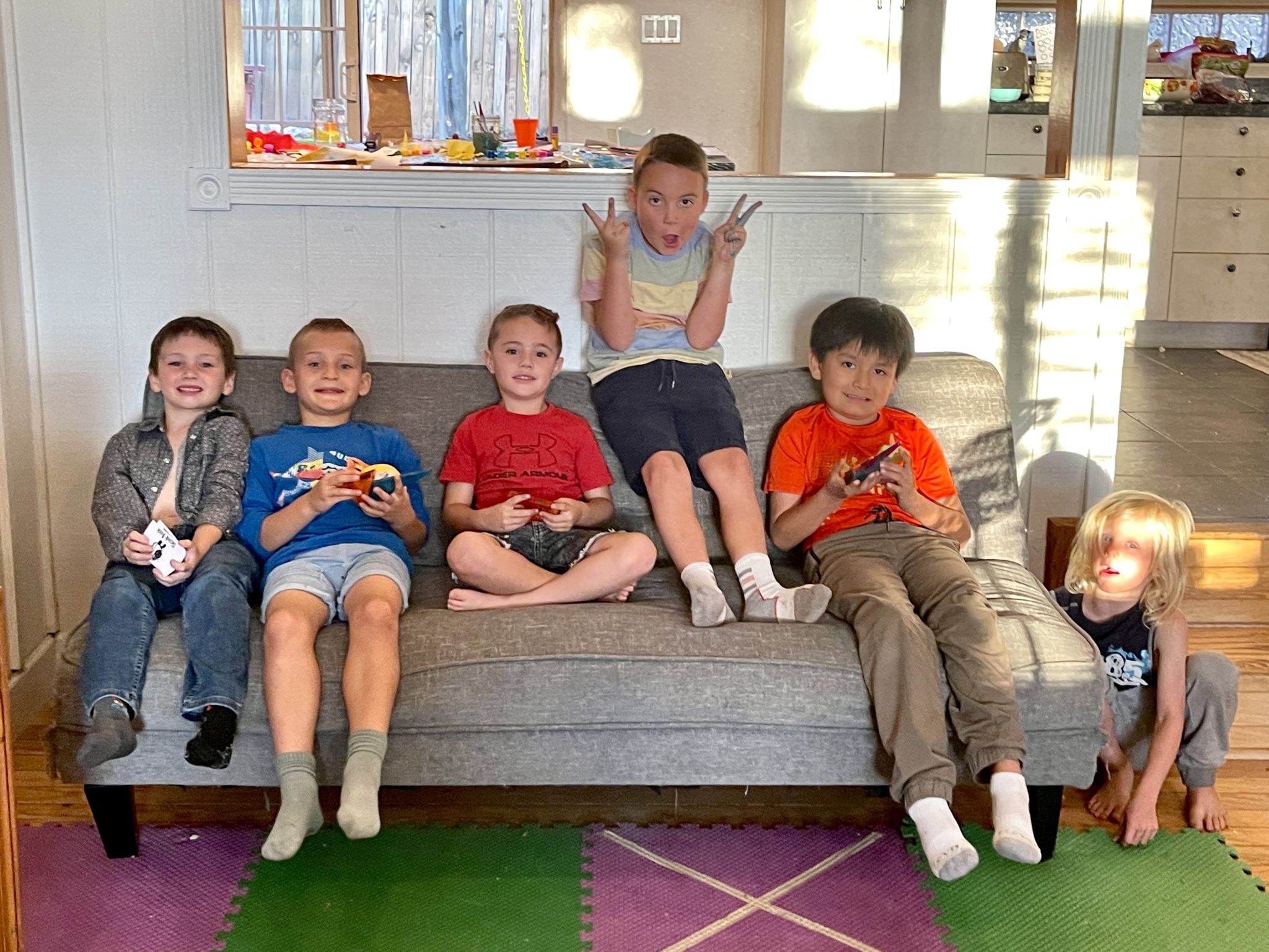 Six boys sitting and posing on a gray couch, some holding small cards, one making peace signs