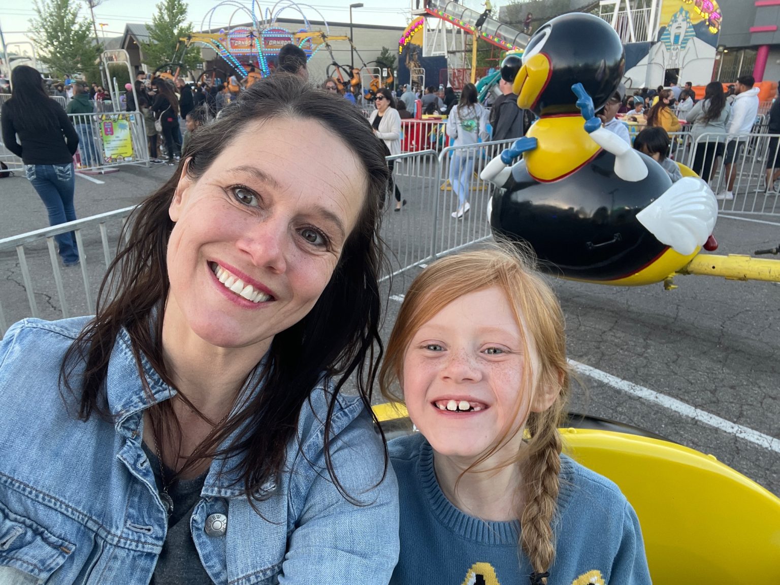 A smiling woman and a young girl seated on a carnival ride, with a ride and crowd behind