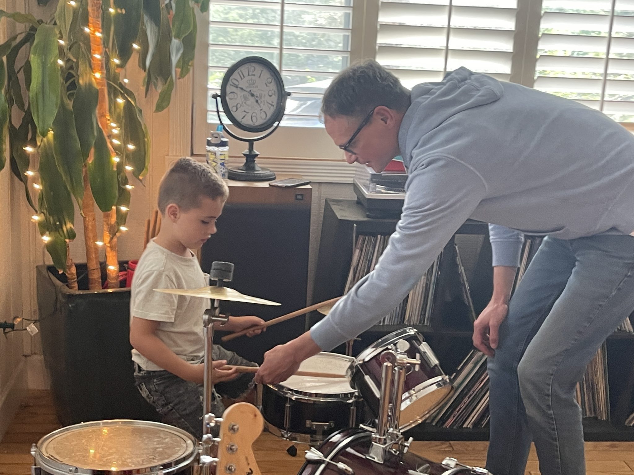 A teacher leaning over to guide a young drummer's sticks at a drum set