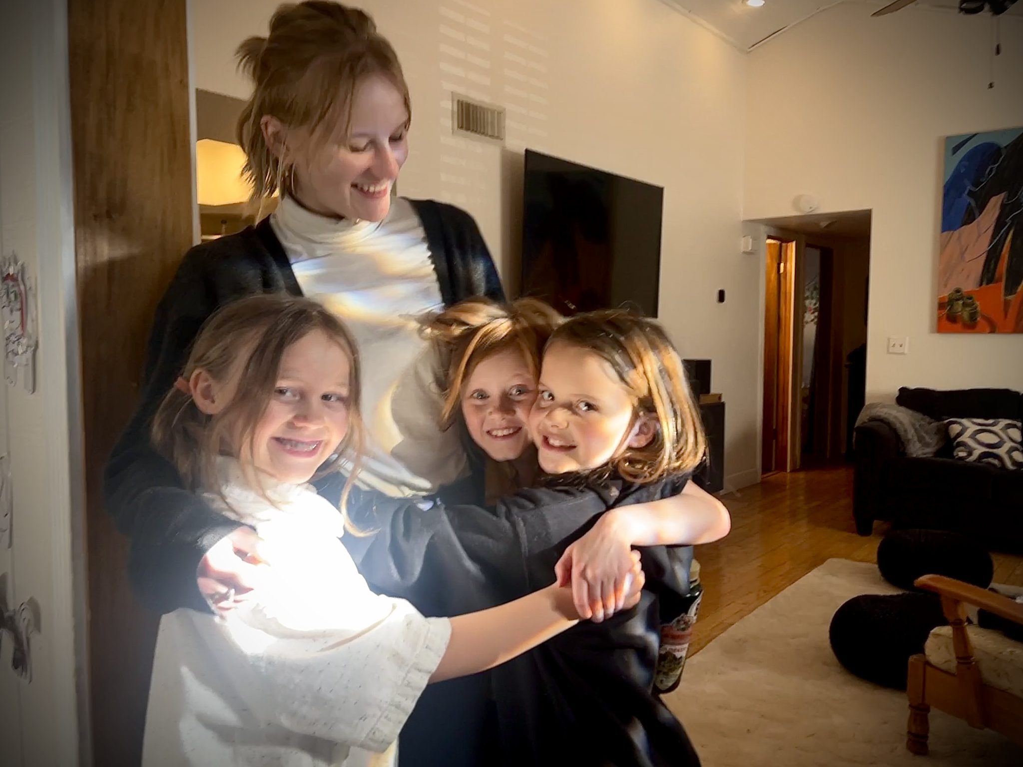 A smiling woman hugged by three smiling kids in a living room