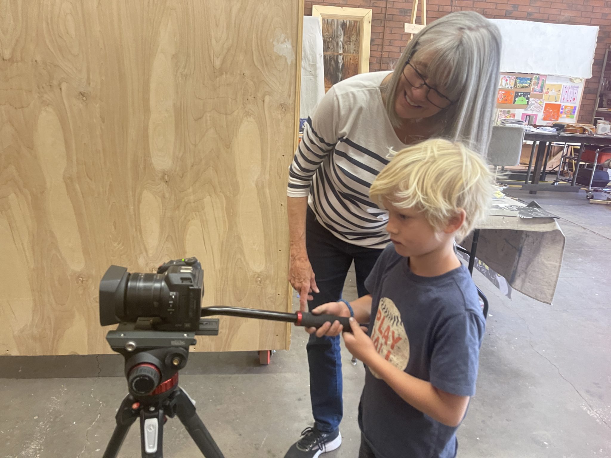 A smiling older woman helping a young child hold a video camera on a tripod