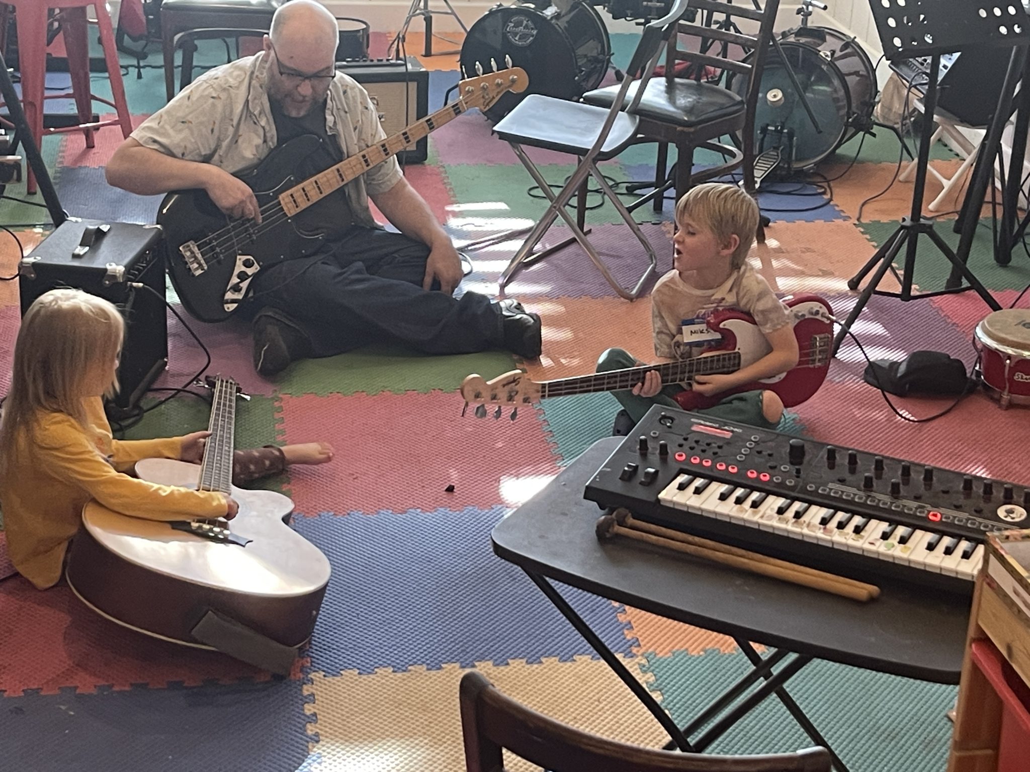 A teacher on bass guitar and two kids with basses on a colorful foam floor, keyboard in front