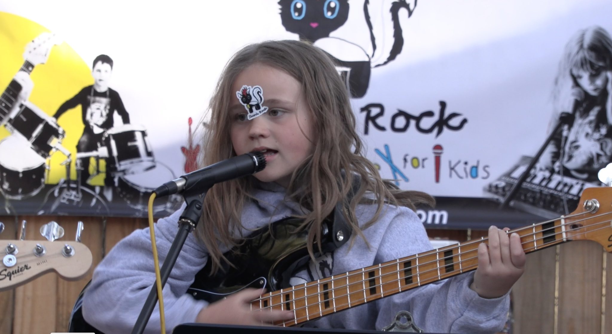 A kid with a skunk sticker on the forehead singing into a microphone and playing bass guitar