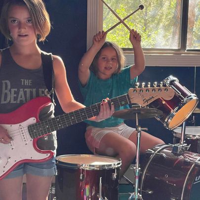 One kid playing a red electric guitar while another holds crossed drumsticks behind a drum set
