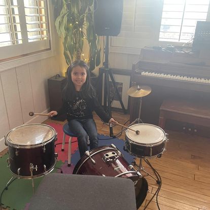A smiling kid sitting at a drum set holding drumsticks in a music room