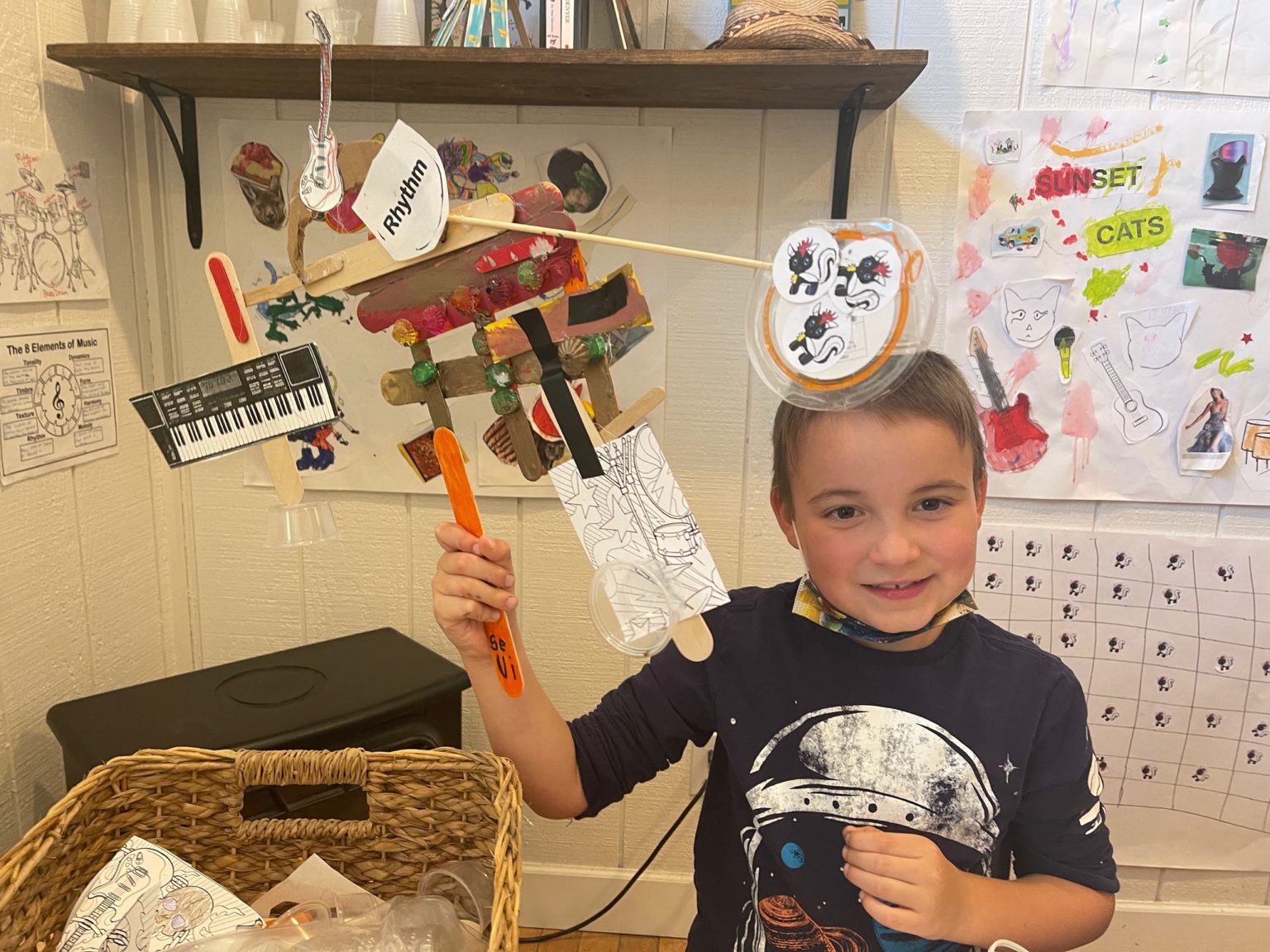 A smiling kid holding up a homemade rhythm craft of craft sticks, paper instruments and skunk stickers