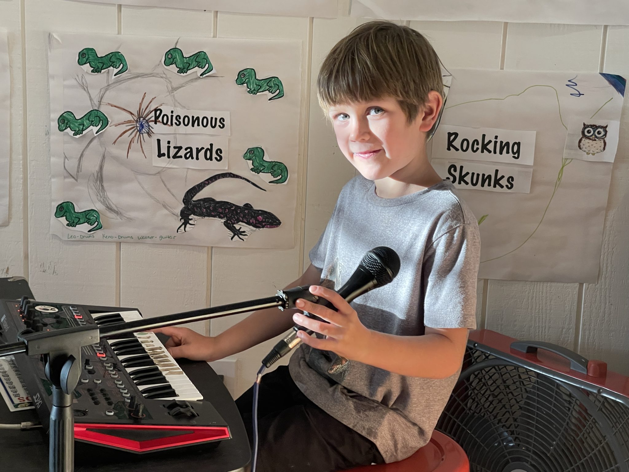 A smiling kid holding a microphone beside a keyboard, in front of posters titled Poisonous Lizards and Rocking Skunks