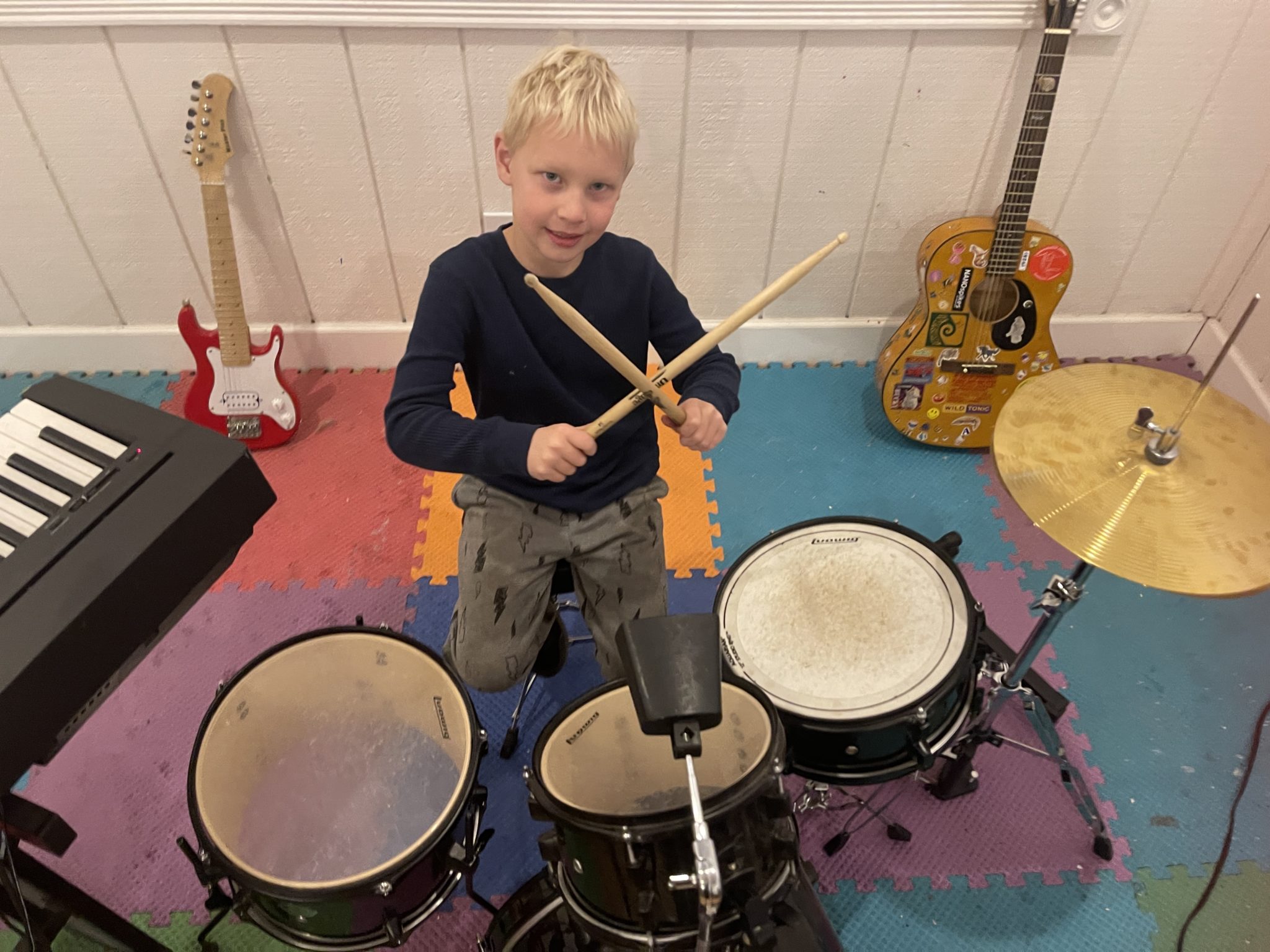 A smiling kid at a drum set holding crossed drumsticks, with guitars and keyboard nearby