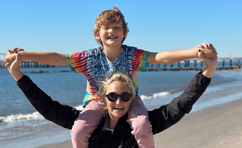 A smiling adult in sunglasses carrying a laughing child on her shoulders at a beach