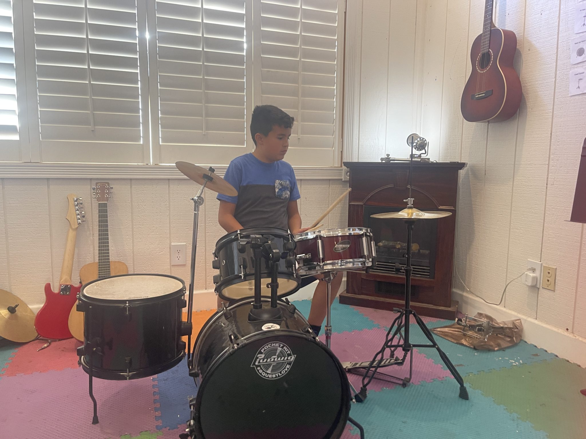 A kid playing a drum set in a bright music room with guitars against the wall
