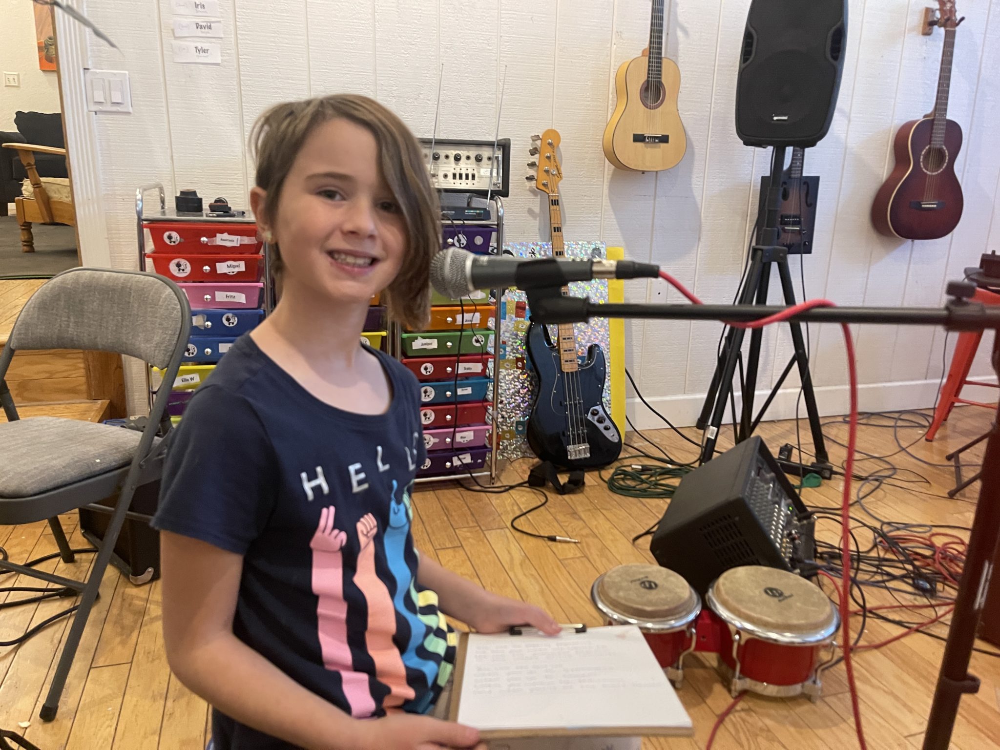 A smiling kid at a microphone holding a clipboard, with bongos and instruments behind