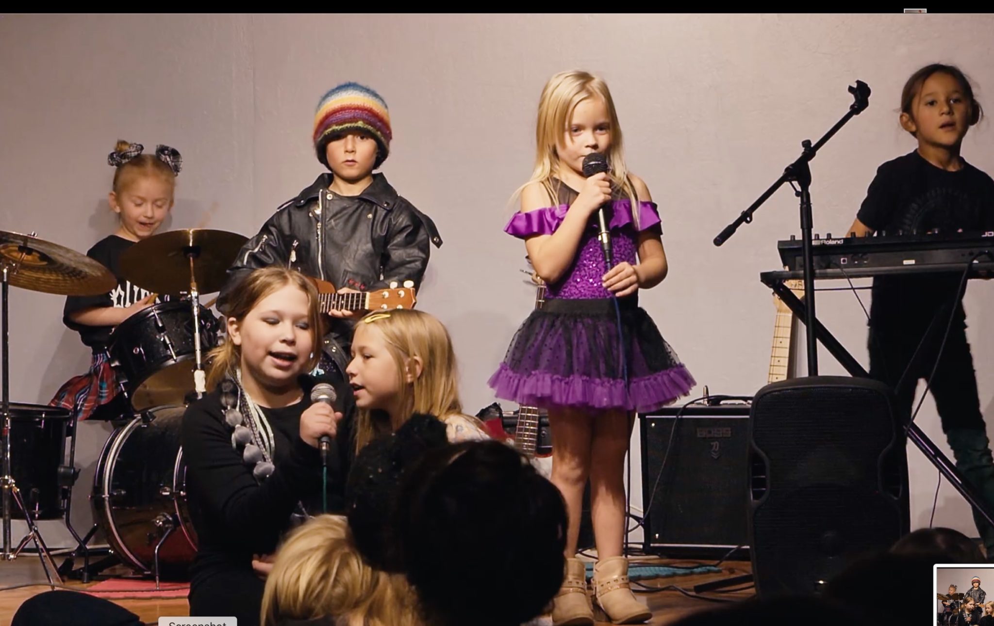 Kids performing on stage, one singing in a purple dress, others on drums, ukulele and keyboard