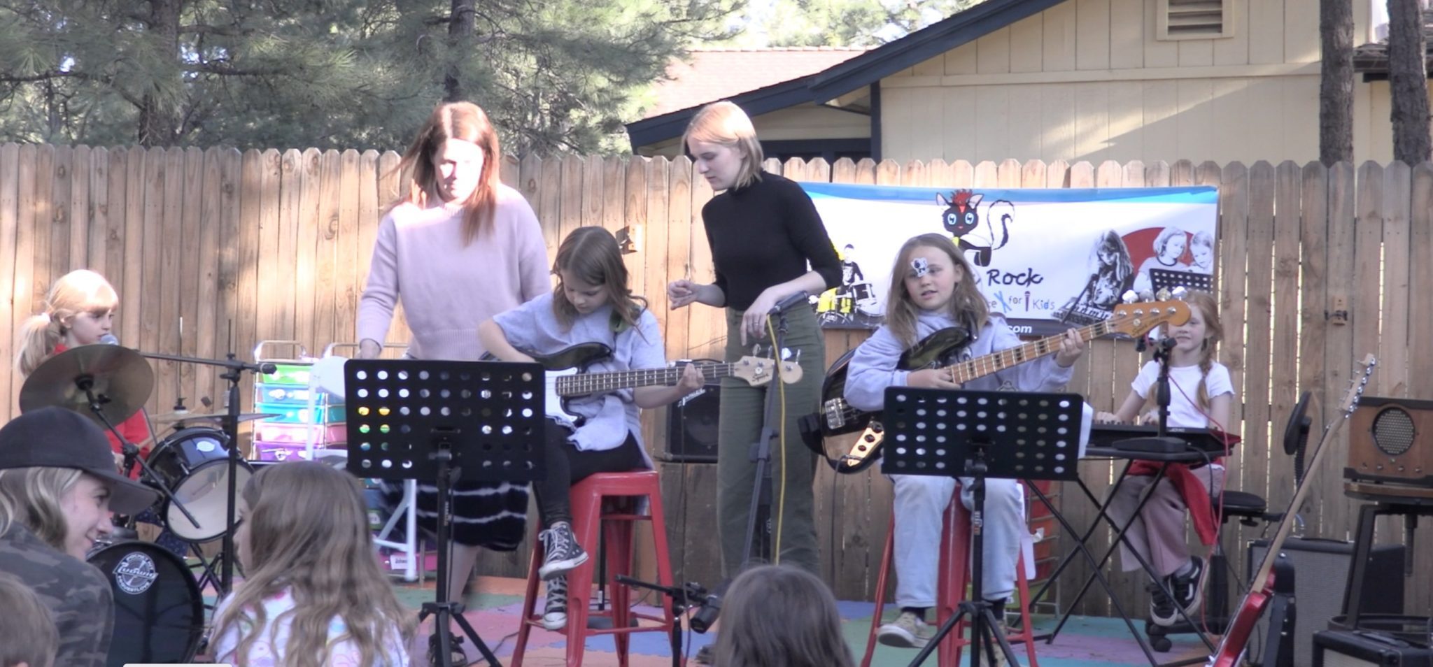 Outdoor show where kids play bass guitars and keyboard while two teachers help, in front of a Skunk Rock banner