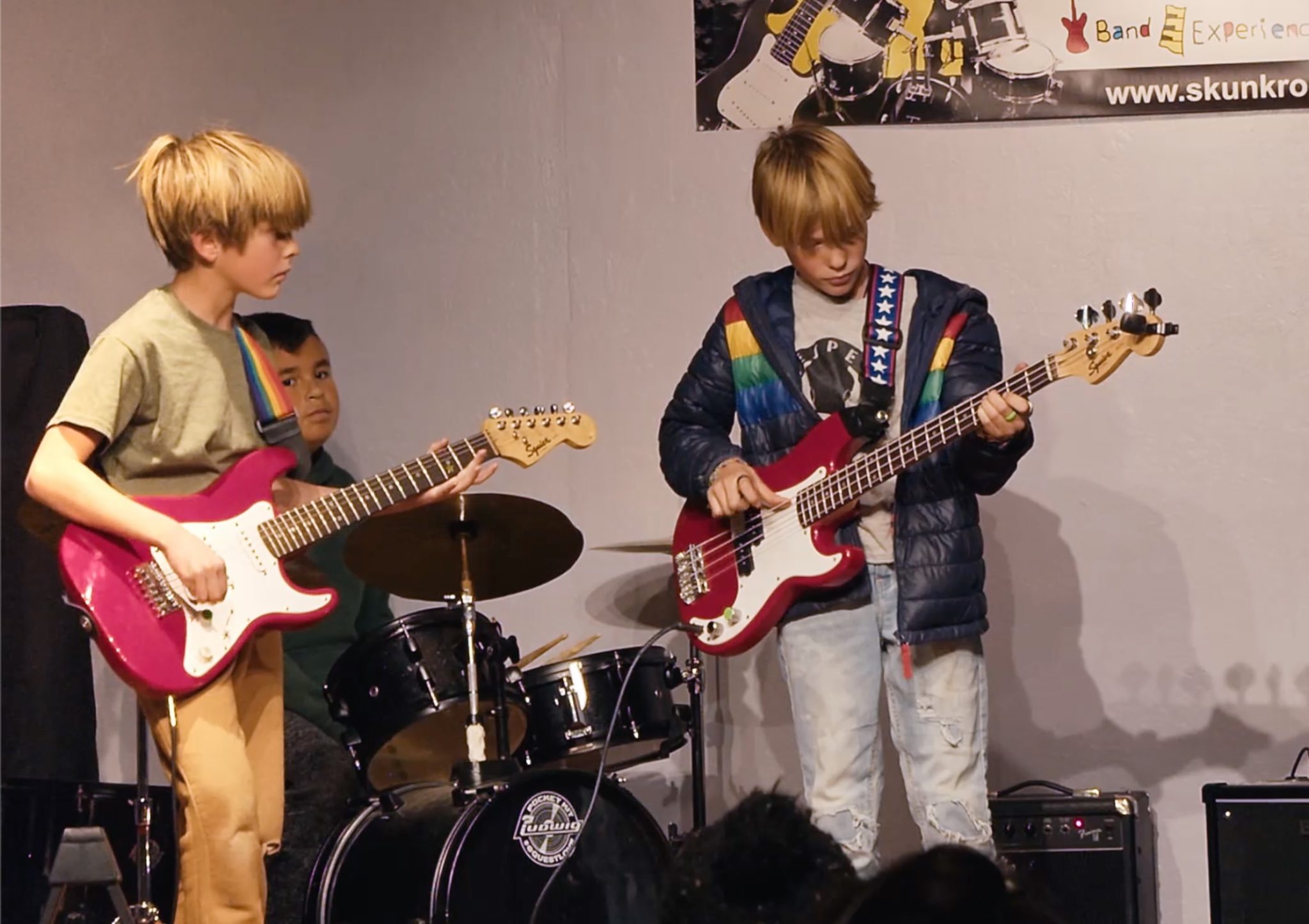 Two kids playing an electric guitar and a red bass on stage, with a drummer behind