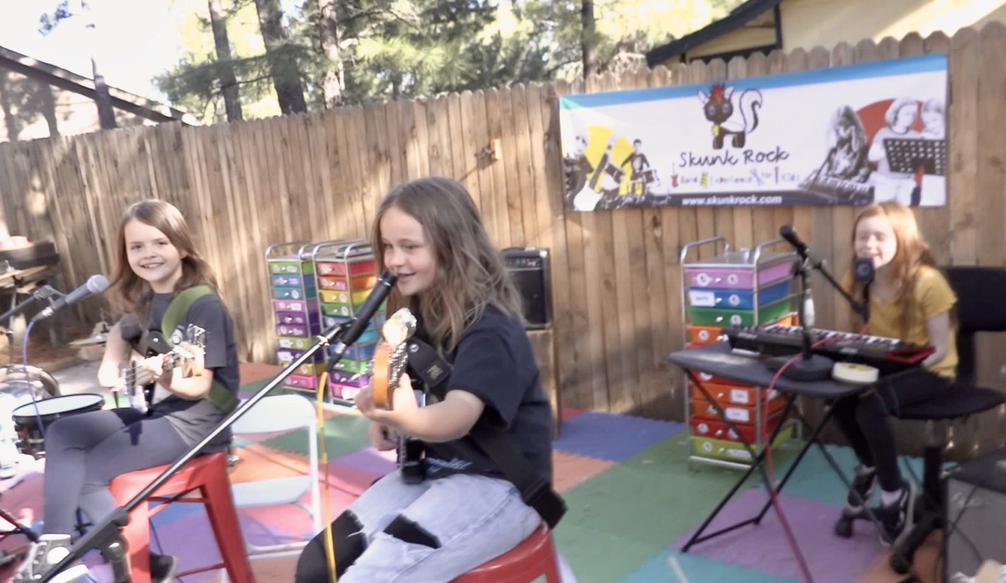Kids playing bass, guitar and keyboard and singing outdoors in front of a Skunk Rock banner