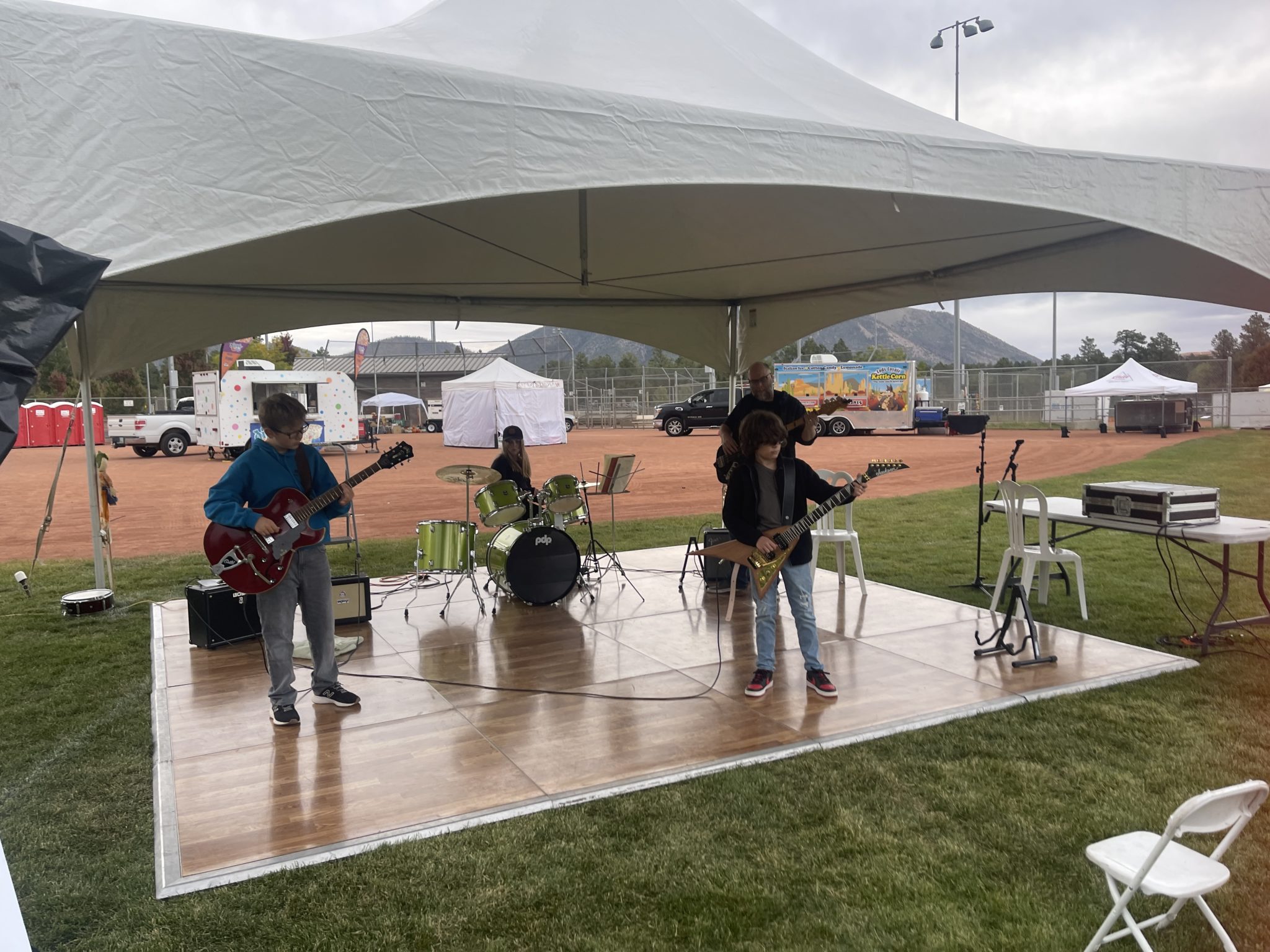 Kids and a teacher playing guitars and drums on a platform under a white tent at an outdoor festival