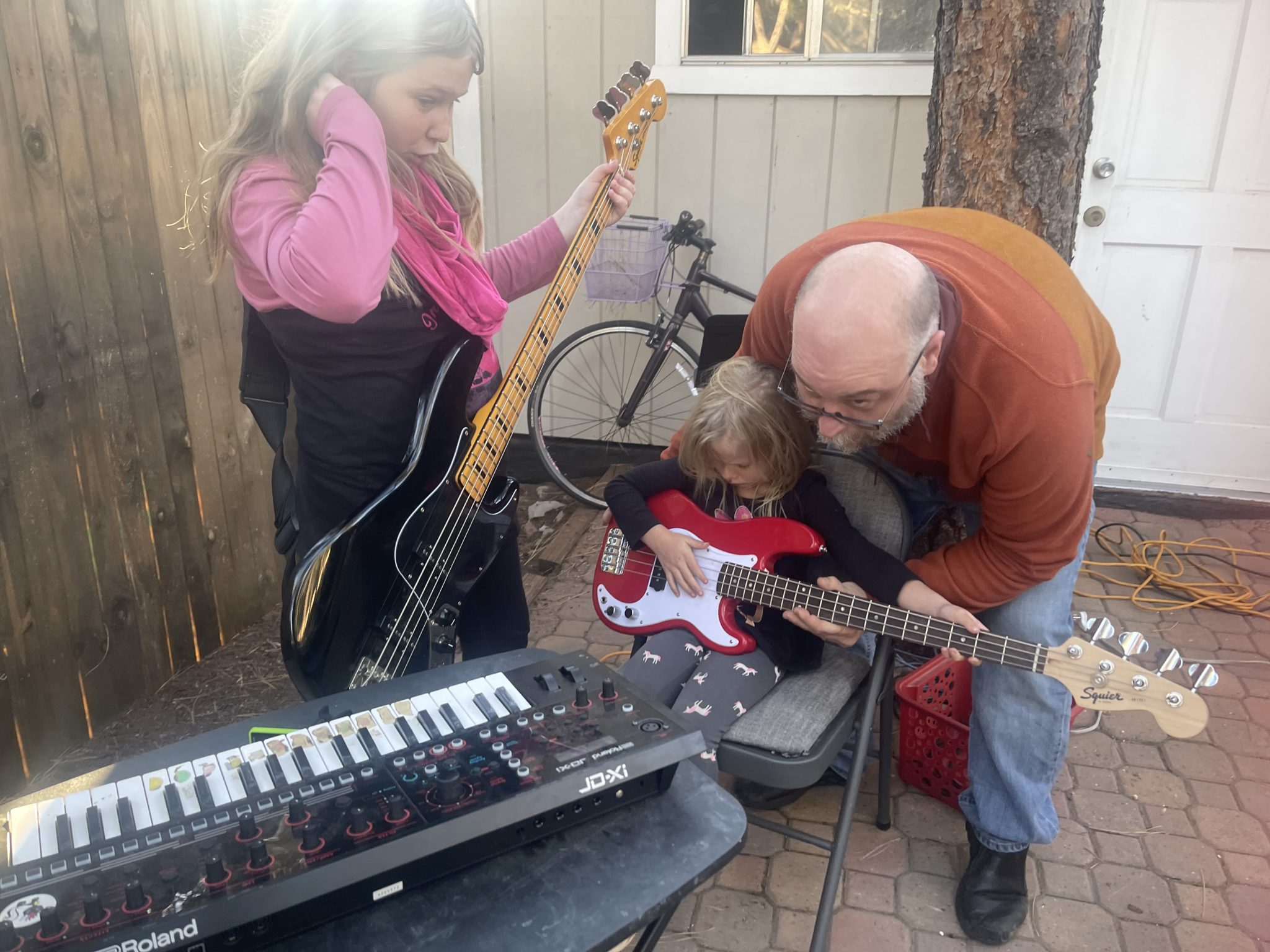 Outdoors, a girl holds a black bass guitar while a teacher helps a small child with a red bass