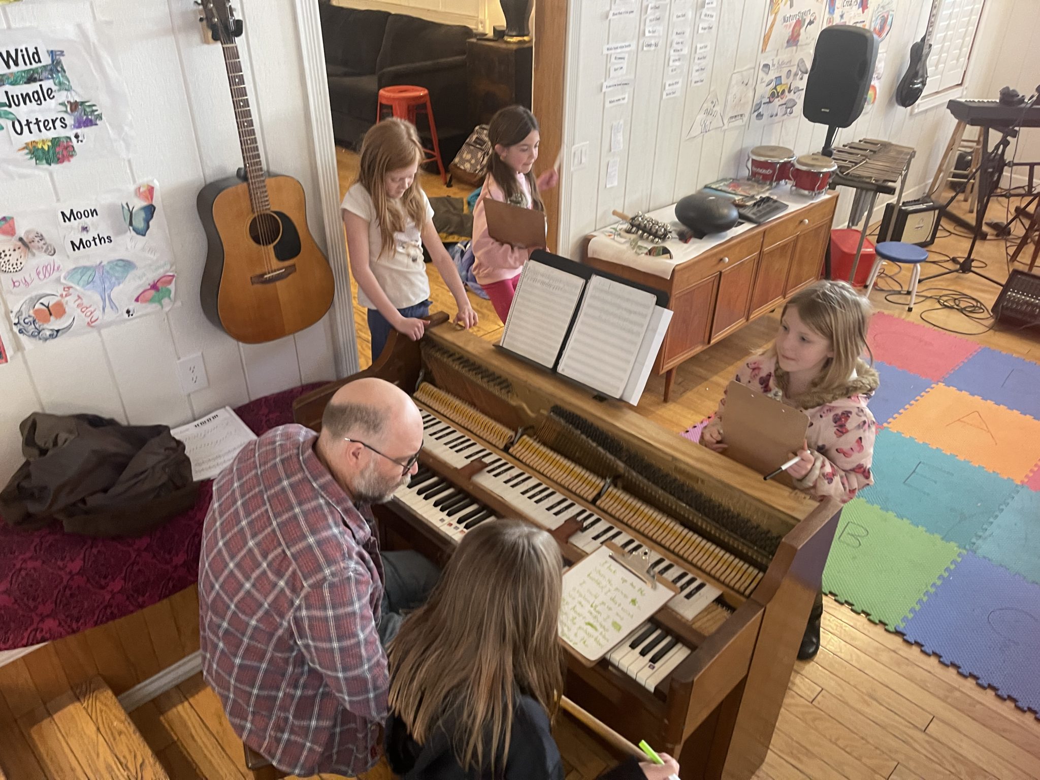 A teacher at an open upright piano with kids holding clipboards, and posters on the wall
