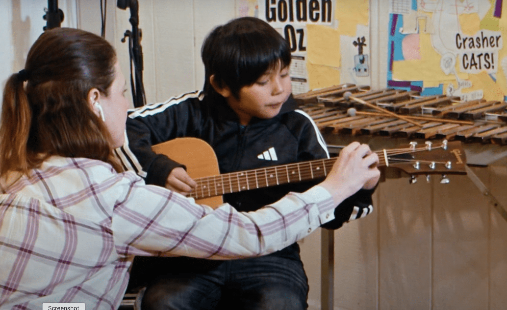 A teacher helping a boy with an acoustic guitar in front of a xylophone and posters