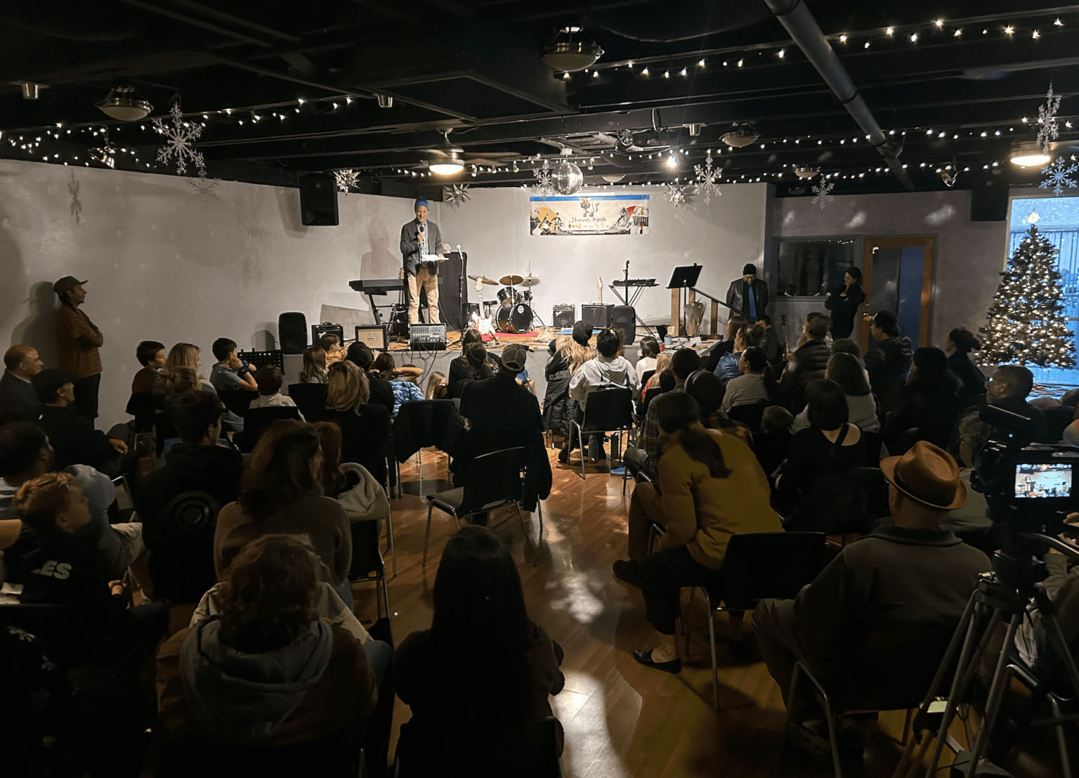 Crowded audience watching a speaker on a decorated stage with a drum set and a Christmas tree