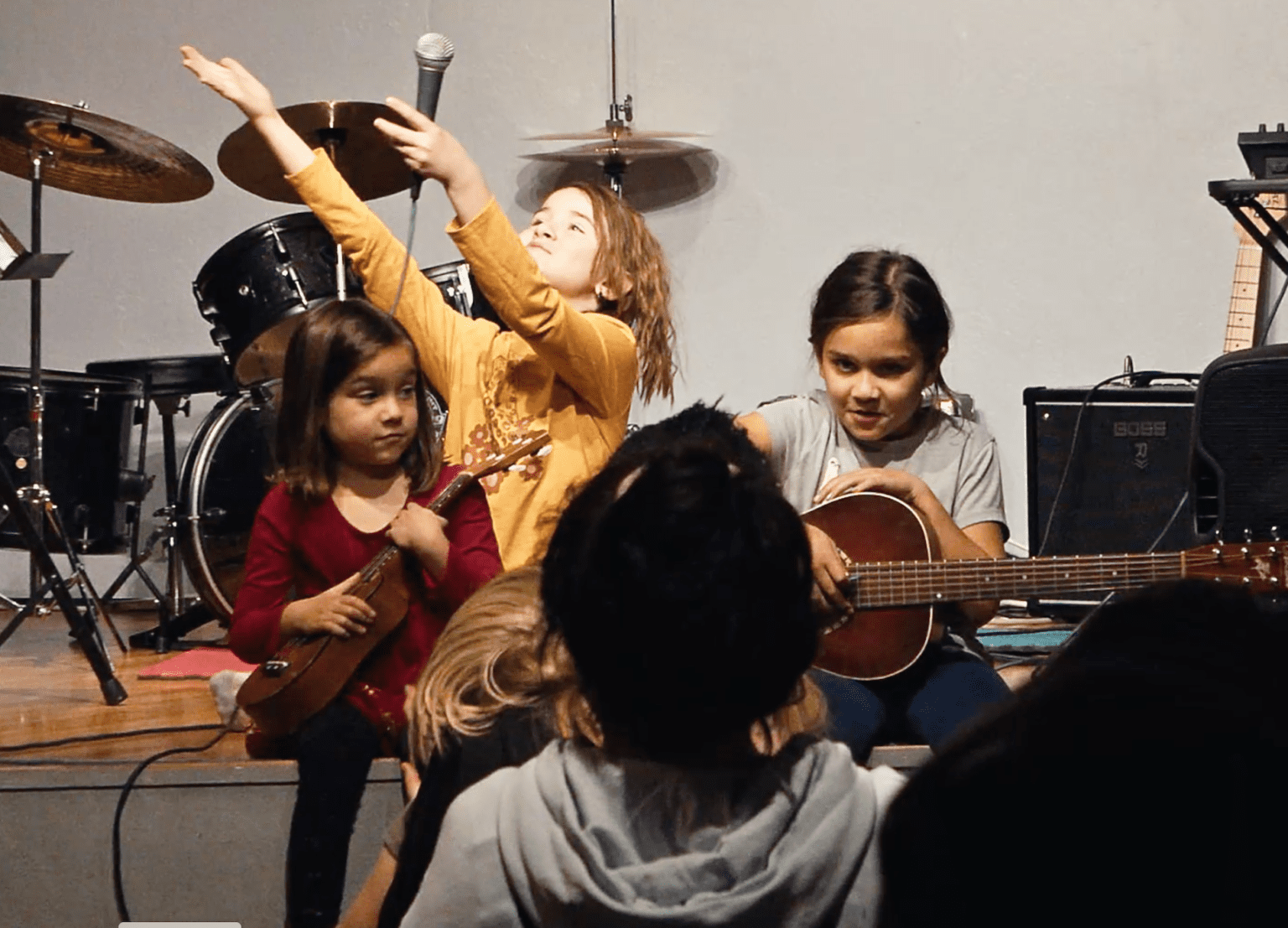 Kids on stage, one raising an arm with a microphone, others holding a ukulele and an acoustic guitar