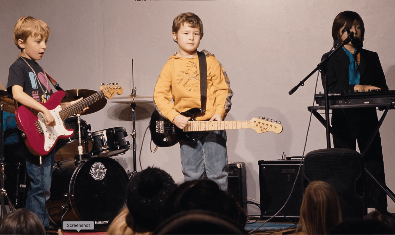 Kids on stage playing two electric guitars and keyboard, with a drum set behind