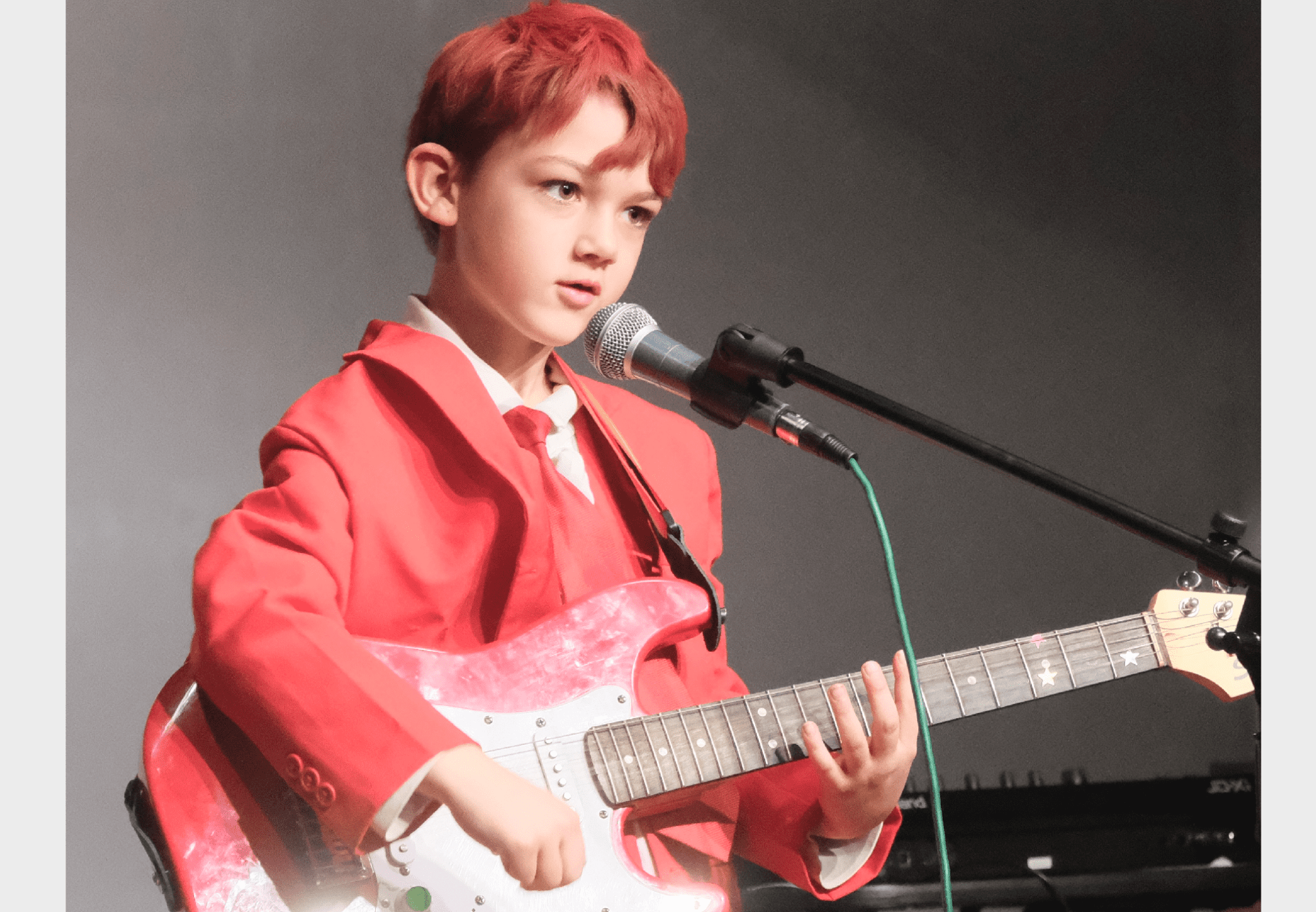 A kid in a red suit and tie playing electric guitar and singing into a microphone