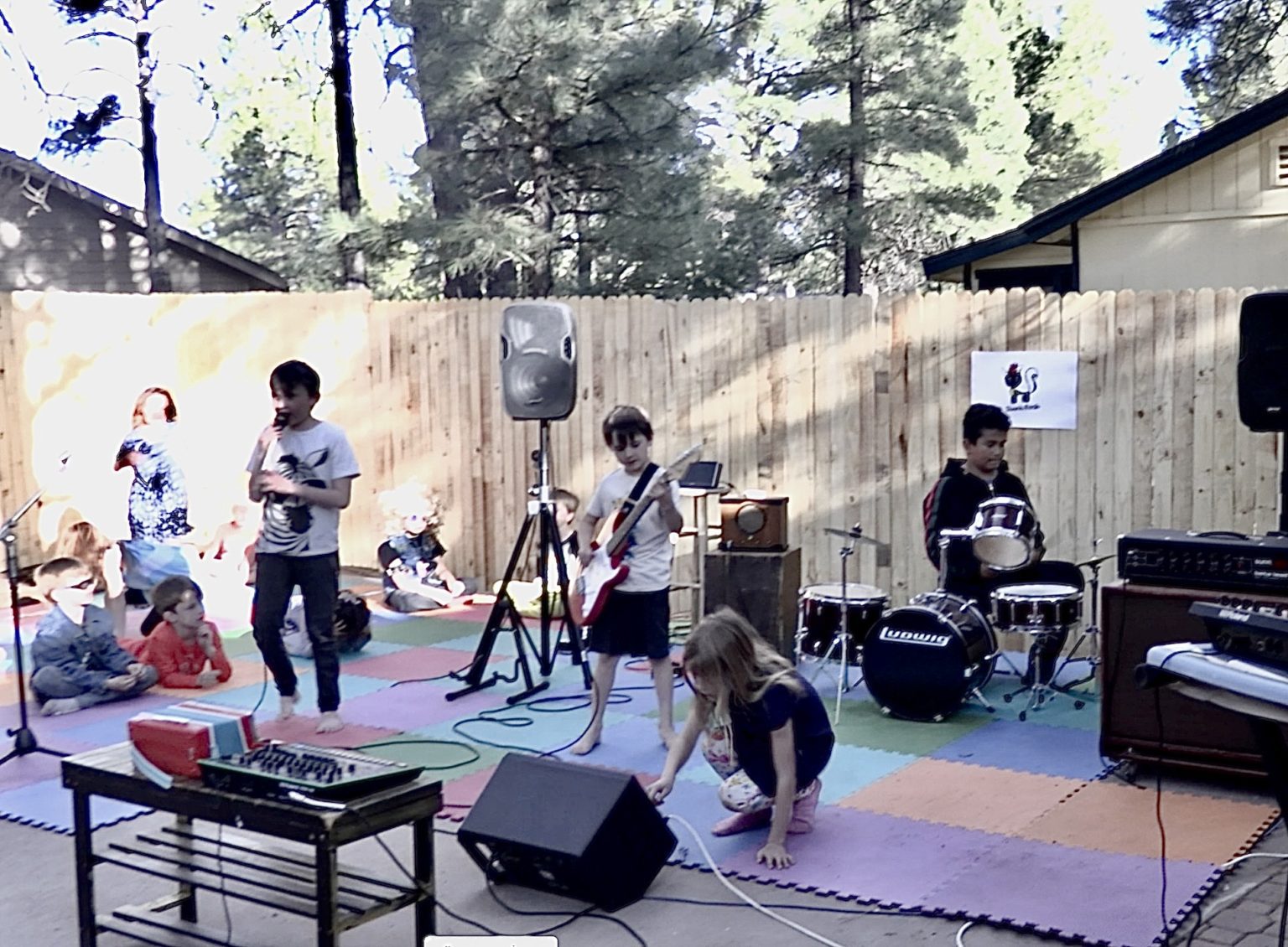 Kids singing, playing guitar and drums at an outdoor show while other kids sit on foam floor tiles